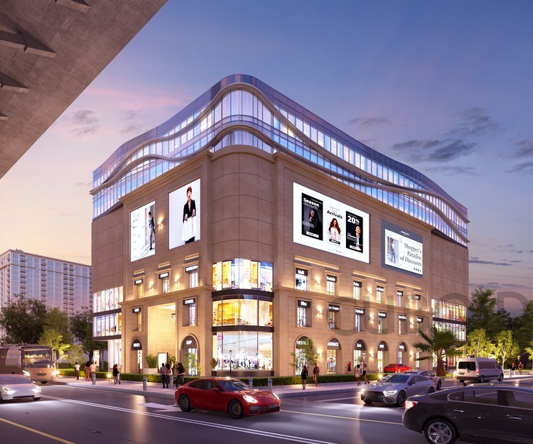 Evening view of a bustling corner shopping mall with illuminated storefronts, large fashion billboards on the façade, curved glass upper floors, and cars moving along the busy street.